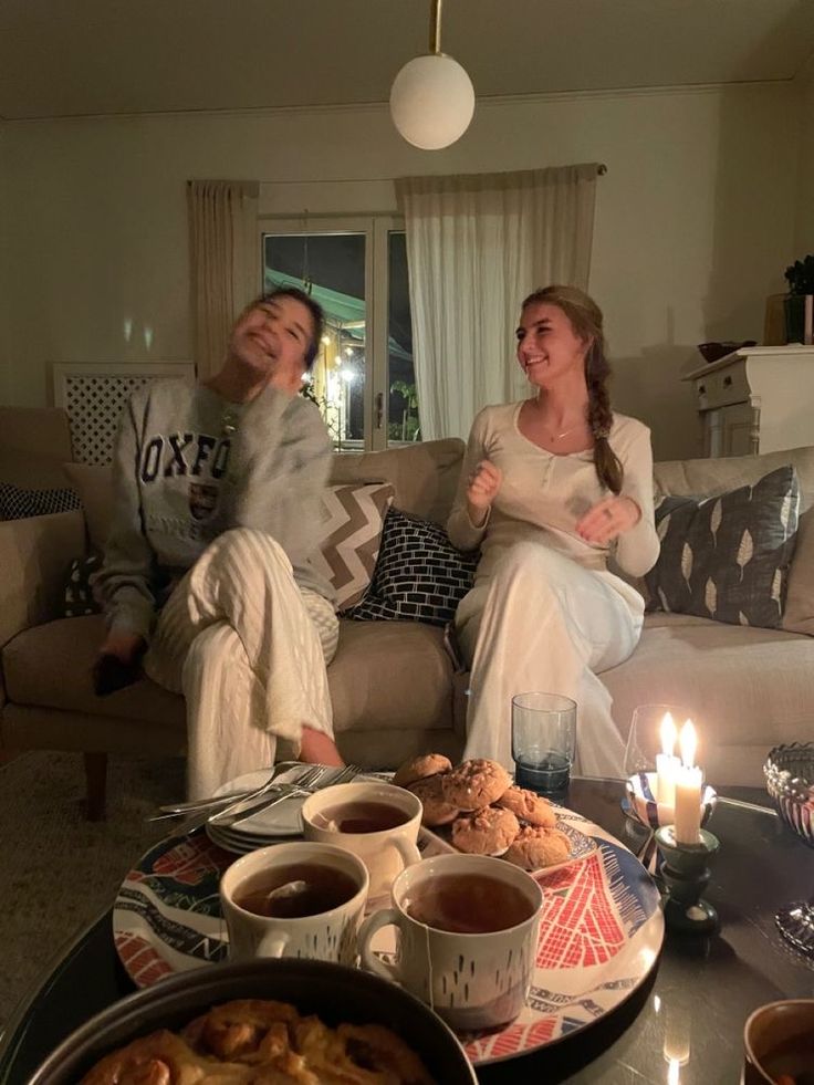 two women sitting on a couch in front of a coffee table with food and candles