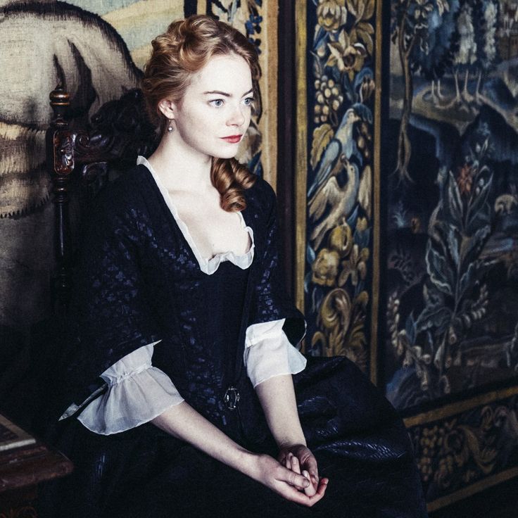 a woman sitting on a chair in front of a wall with ornate paintings behind her