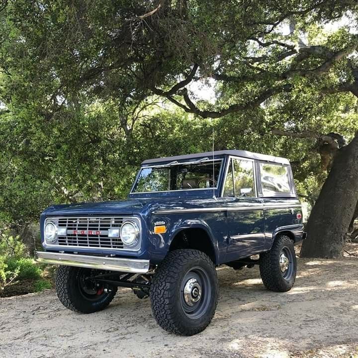 an old blue truck is parked in the dirt near some trees and bushes on a sunny day