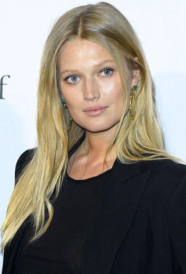 a woman with long blonde hair wearing a black top and gold hoop earrings, standing in front of a white background