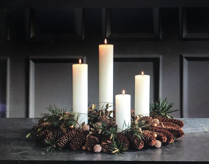 four white candles sitting on top of a table next to pine cones