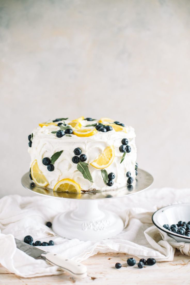 a white cake with blueberries and lemons on it sitting next to a bowl of blueberries