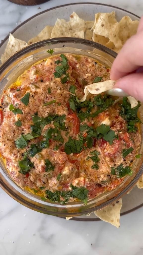 a person dipping tortilla chips into a glass bowl filled with sauce and herbs