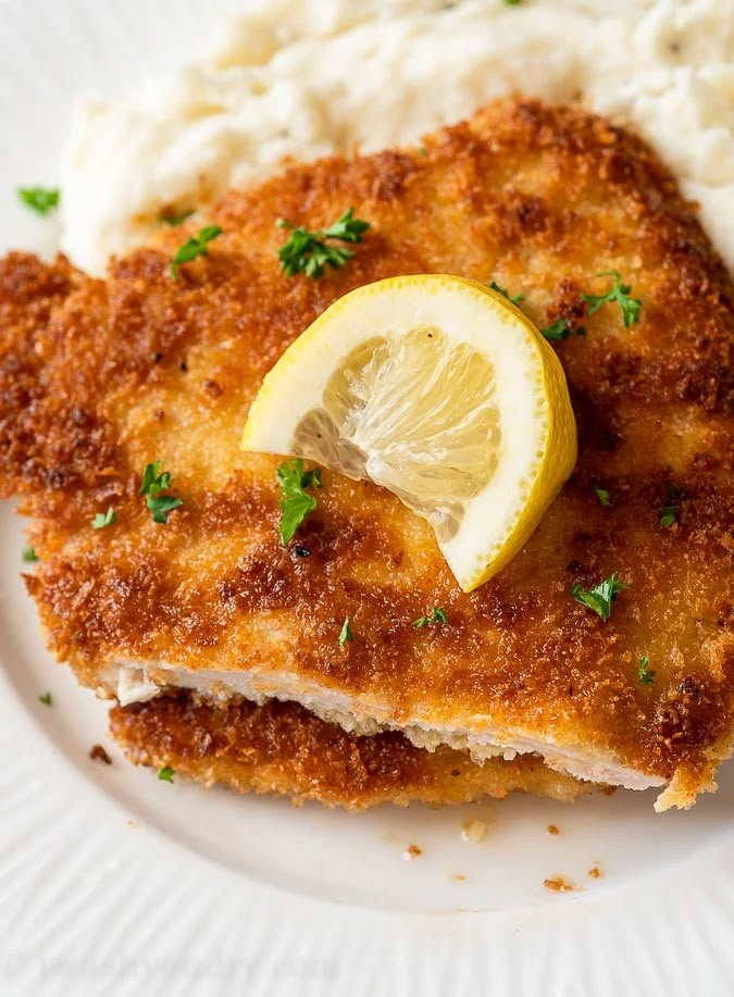 a white plate topped with chicken and mashed potatoes next to a lemon wedge on top of mashed potatoes