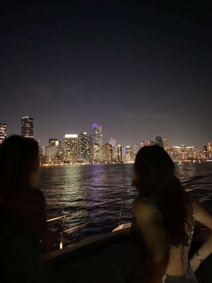 two women are sitting on a boat looking at the city lights from across the water