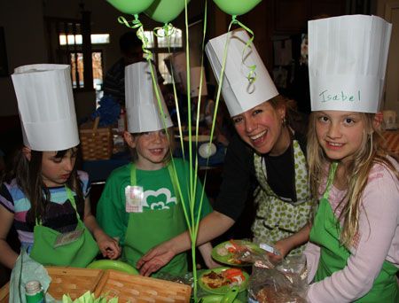 four children wearing chefs hats and aprons around a table with food in front of them