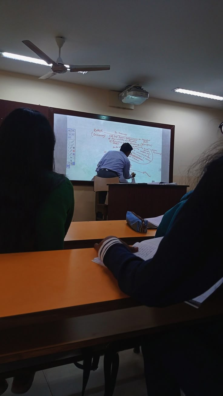 three people are sitting at desks in front of a projector screen and one person is writing on the board