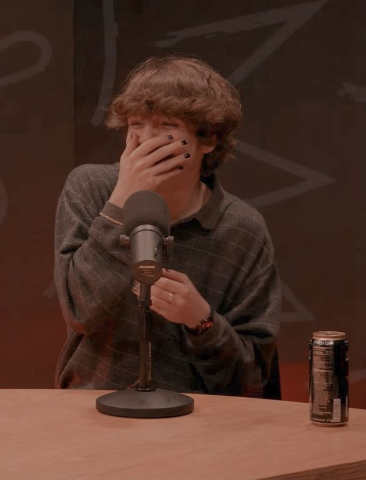 a boy covering his face while sitting at a table with a microphone in front of him