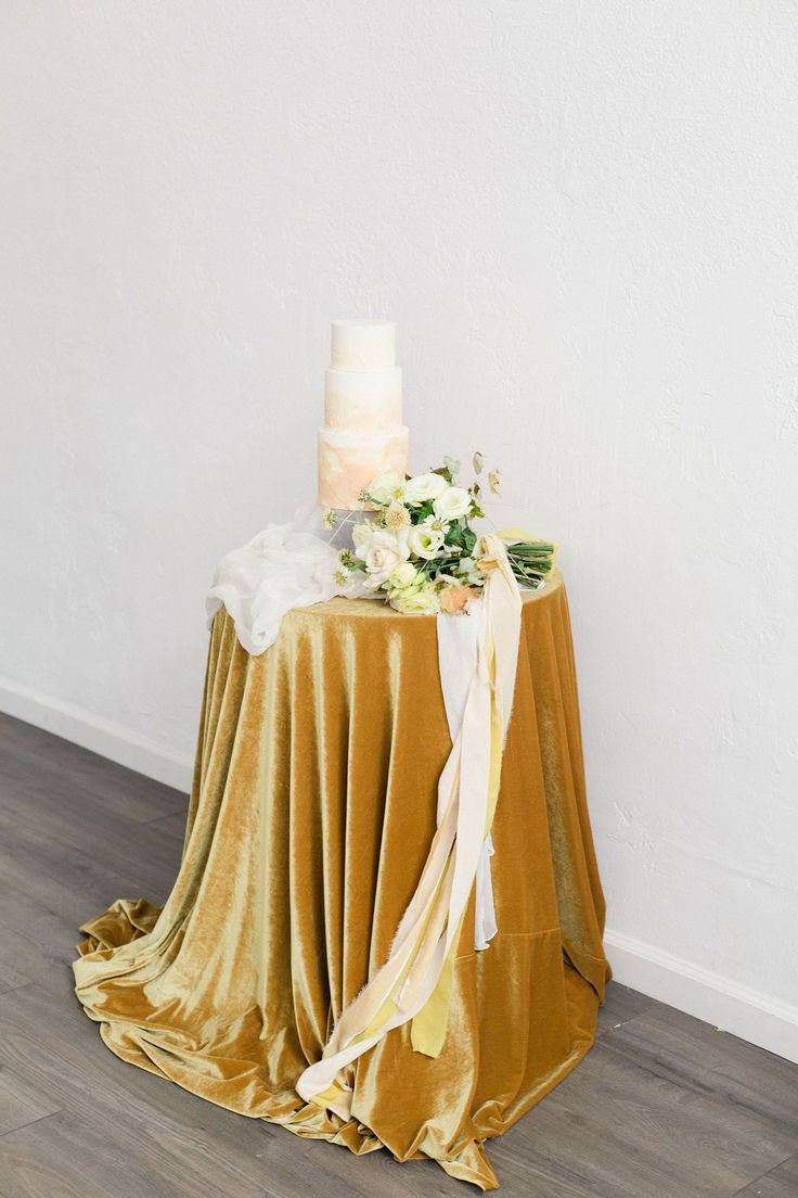 a wedding cake on top of a gold table cloth with white flowers and greenery