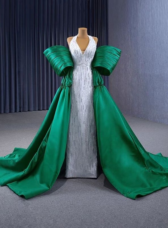 Regal and captivating, this prom dress is designed for those who wish to exude elegance and confidence. The halter neckline and form-fitting silver bodice are embellished with dazzling beadwork, creating a sophisticated and glamorous look. The standout feature of this gown is the dramatic emerald green overskirt, adorned with oversized bow accents that add a unique and stylish touch. The voluminous skirt flows into a majestic train, enhancing the dress's regal silhouette and ensuring you make a memorable entrance. Imagine gliding across the dance floor, the fabric shimmering with every step, capturing the admiration of everyone around you. This dress is ideal for those who want to combine contemporary fashion with classic beauty. Let this stunning gown transform your prom night into a magi Green Satin Prom Dress, Quinceanera Dresses Green, Red Quinceanera Dress, Champagne Quinceanera Dresses, White Quinceanera Dresses, Purple Quinceanera Dresses, Quinceanera Dresses Gold, Black Quinceanera Dresses, Quinceanera Dresses Pink