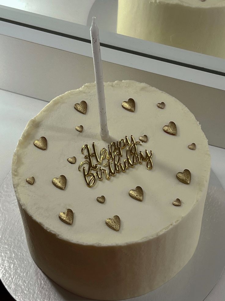 a white cake with gold hearts on it and a happy birthday candle in the middle