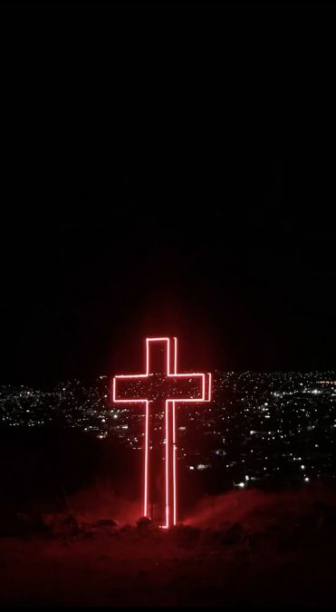 a lighted cross on top of a hill in the night sky with city lights behind it