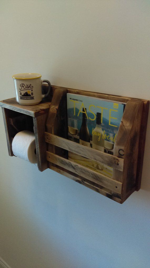 a wooden shelf with magazines and coffee mugs on it next to a wall mounted toilet paper dispenser