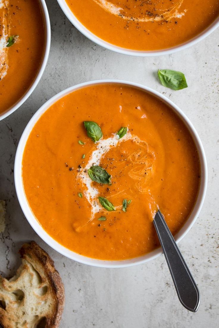 three bowls of tomato soup with basil and parmesan cheese on the top, next to bread