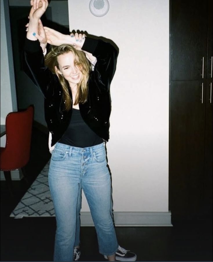 a woman in jeans and black jacket holding her arms up with both hands while standing next to a white wall