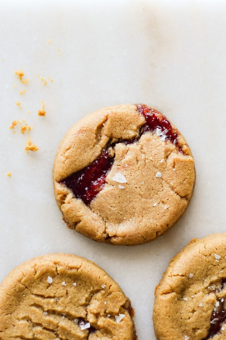 three peanut butter and jelly cookies on a white surface with crumbs around them