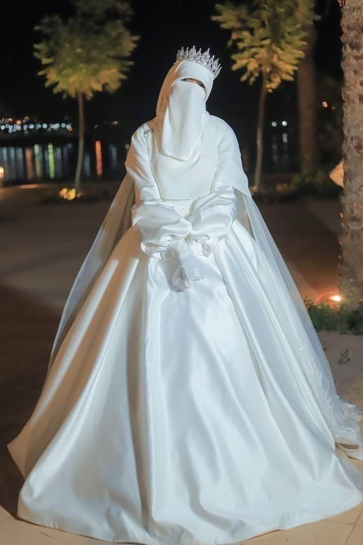 a woman in a white wedding gown and veil is standing on the sidewalk at night