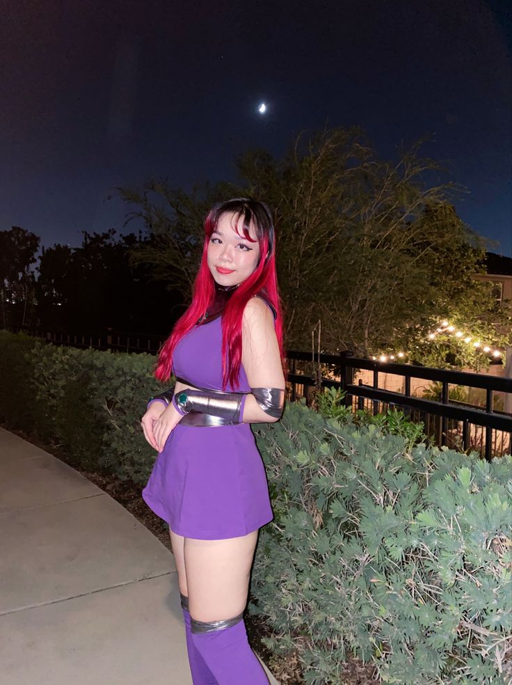 a woman with red hair and purple dress posing for the camera in front of bushes