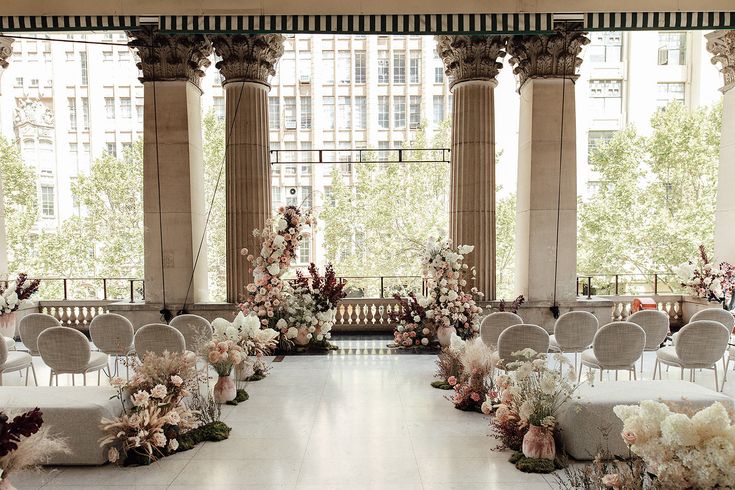 an outdoor ceremony setup with white chairs and floral arrangements on the floor in front of large windows