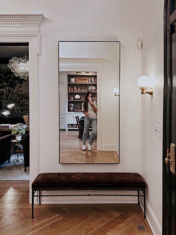 a woman standing in front of a mirror on the wall next to a wooden bench