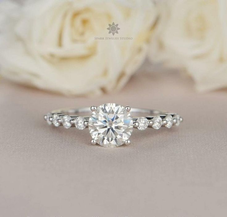 a close up view of a diamond engagement ring with white roses in the foreground