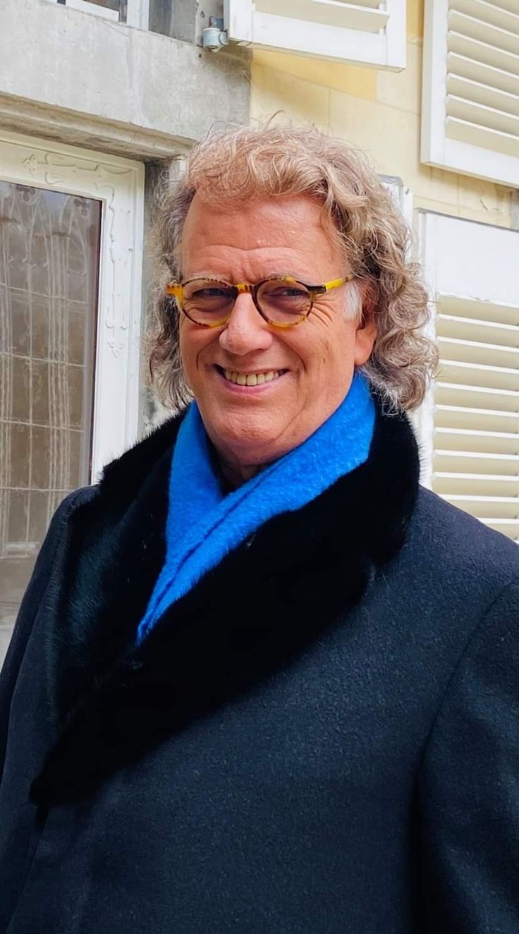 a man with glasses and a blue scarf standing in front of a building smiling at the camera