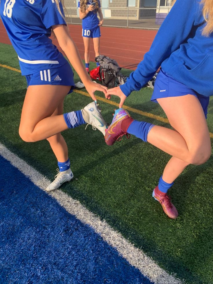 two girls in blue uniforms holding hands on a soccer field with one girl wearing pink shoes