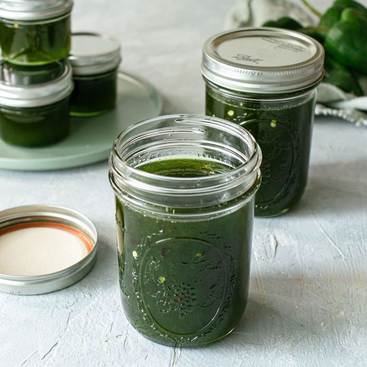 three jars filled with green liquid sitting on top of a white table next to some vegetables