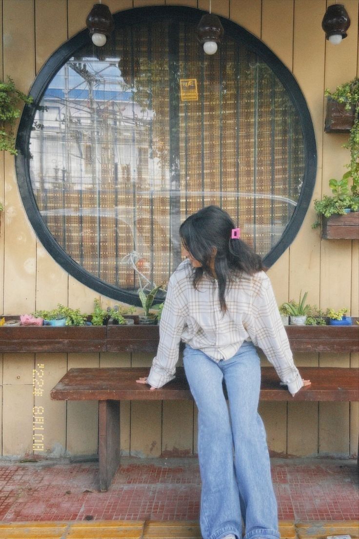 a woman sitting on top of a wooden bench next to a wall filled with potted plants