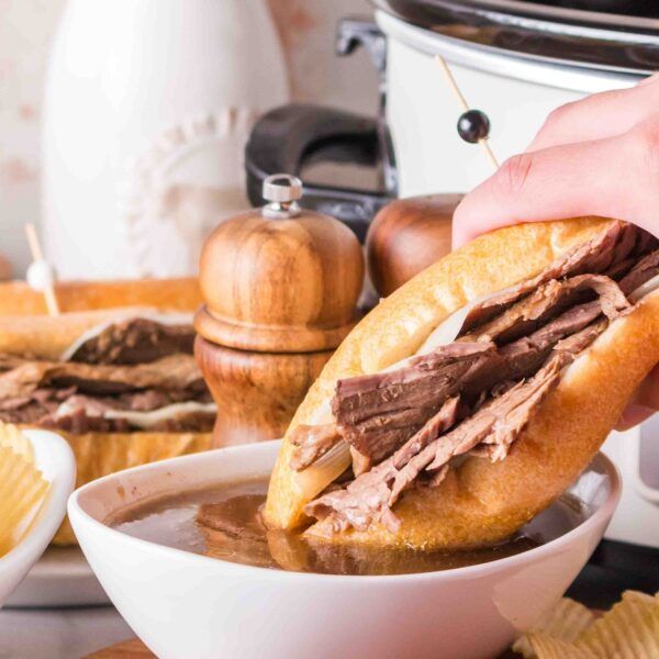 a person is dipping some food into a bowl with chips on the side and an instant pot roast beef sandwich in the background