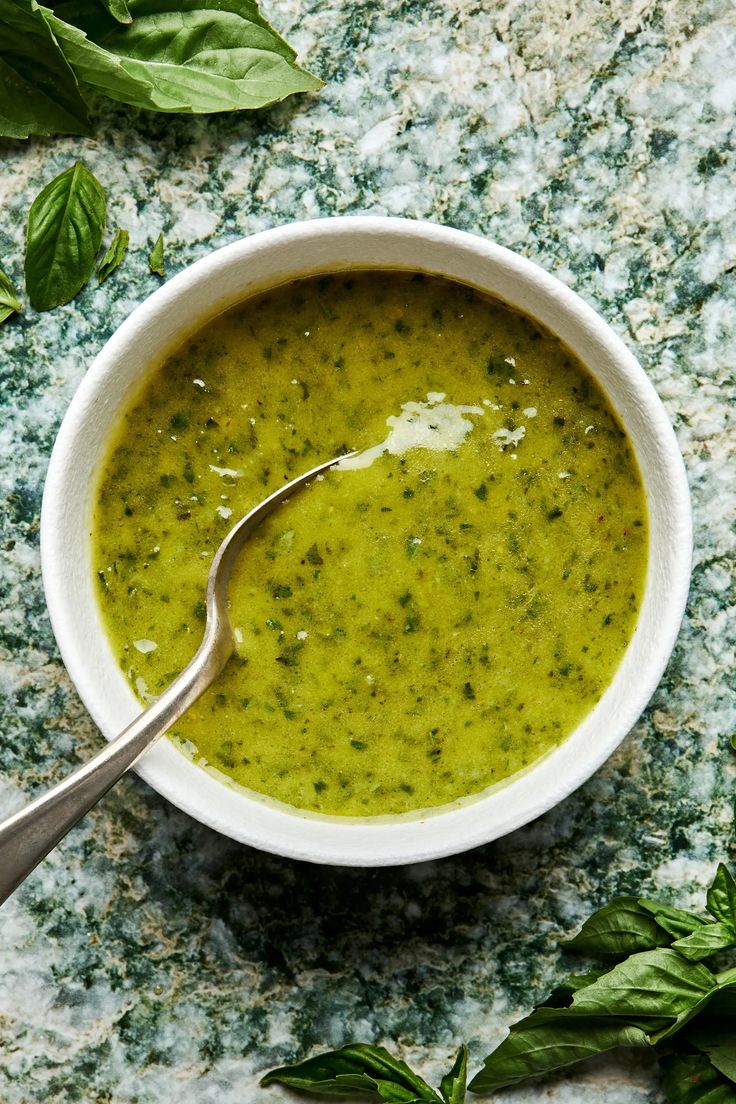 a white bowl filled with pesto sauce on top of a marble counter next to basil leaves