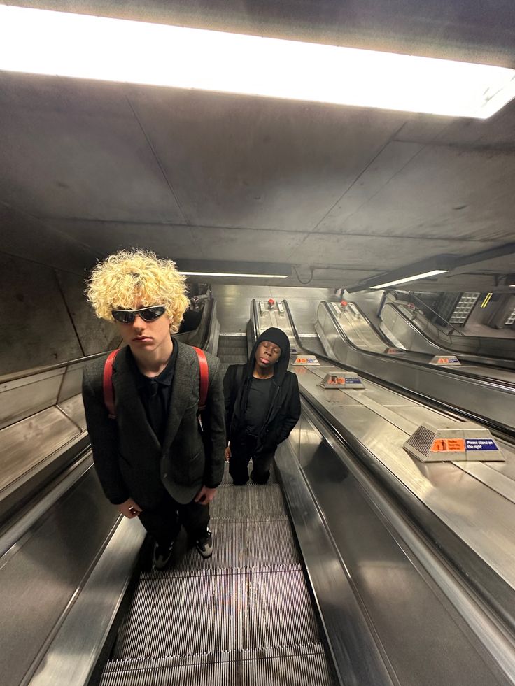 two young men standing on an escalator in a subway station, one wearing sunglasses and the other with blonde hair