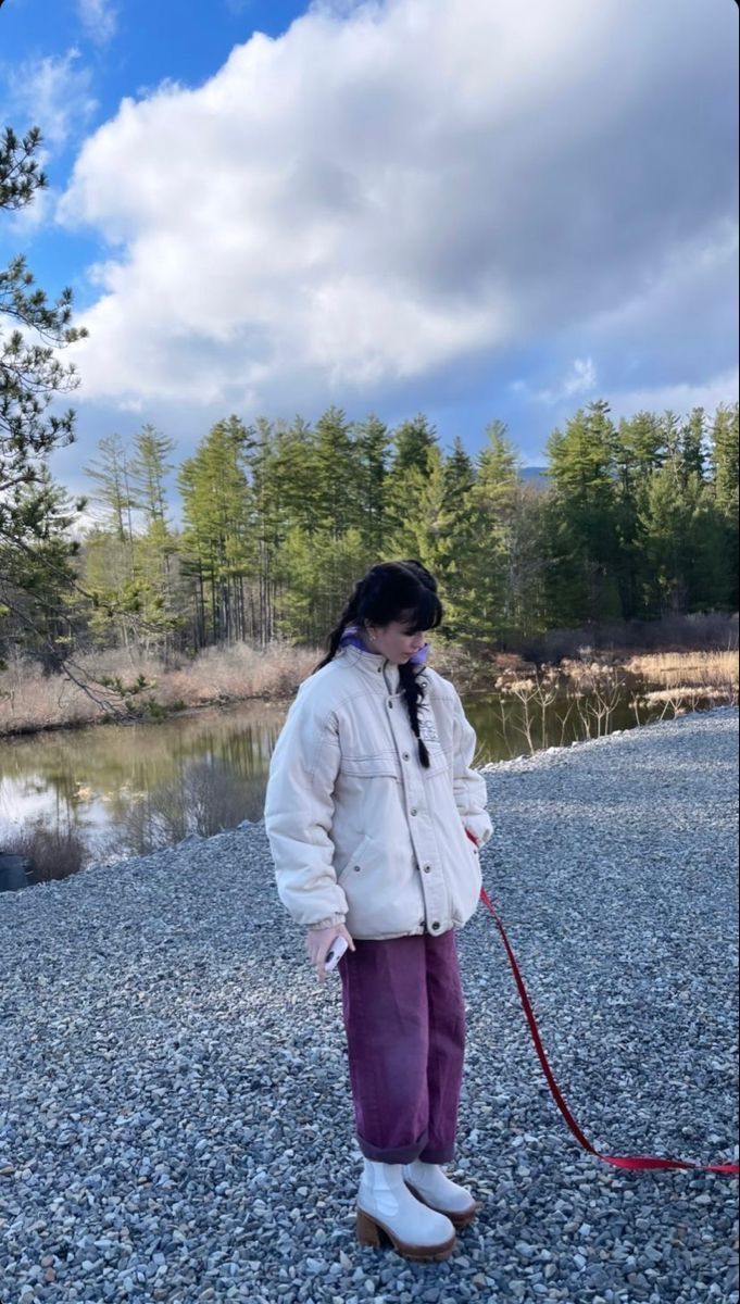 a woman in white jacket holding a red leash on gravel ground next to water and trees