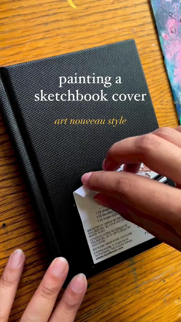 someone is painting a sketchbook cover on a table with their hands holding the book