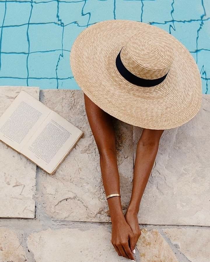 a woman sitting on the edge of a swimming pool wearing a straw hat and reading a book