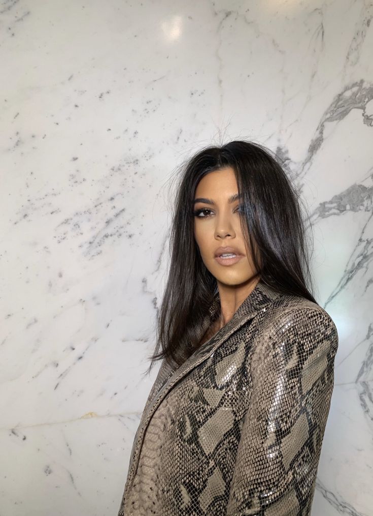 a woman with long hair standing in front of a marble wall wearing a snake skin jacket