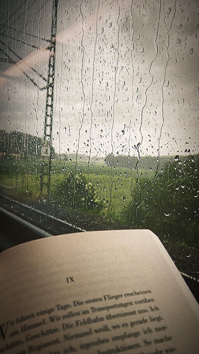 an open book sitting on top of a table next to a window covered in rain