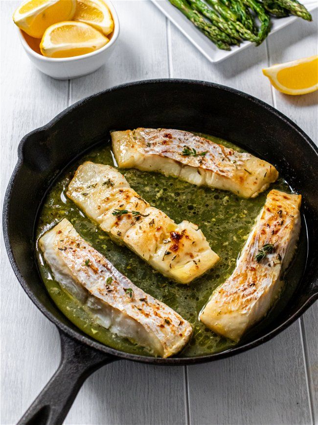 some fish is cooking in a skillet with asparagus and lemons on the side