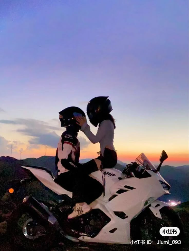 two people sitting on a motorcycle in the mountains at sunset or dawn with one person touching the other's face