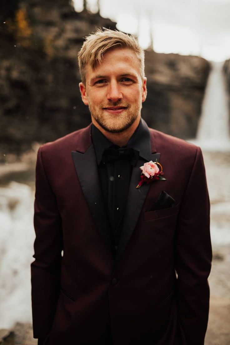 a man in a tuxedo standing next to a waterfall wearing a boutonniere