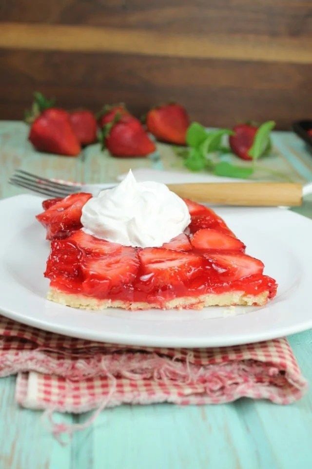 a slice of strawberry pie on a plate with whipped cream and strawberries in the background