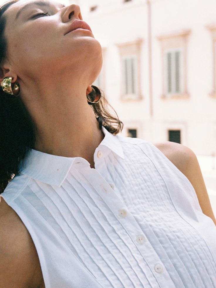 a woman in white shirt and gold earrings looking up at the sky with buildings behind her