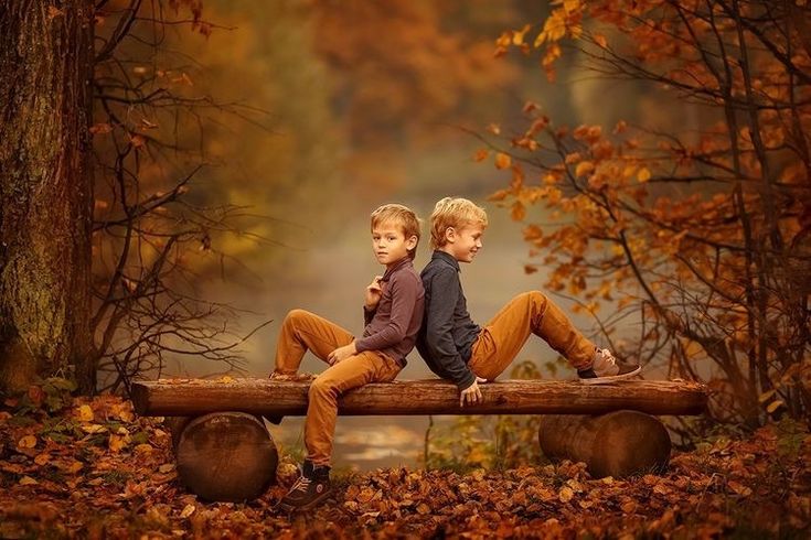 two young boys sitting on a log in the woods with autumn leaves around them and trees behind them