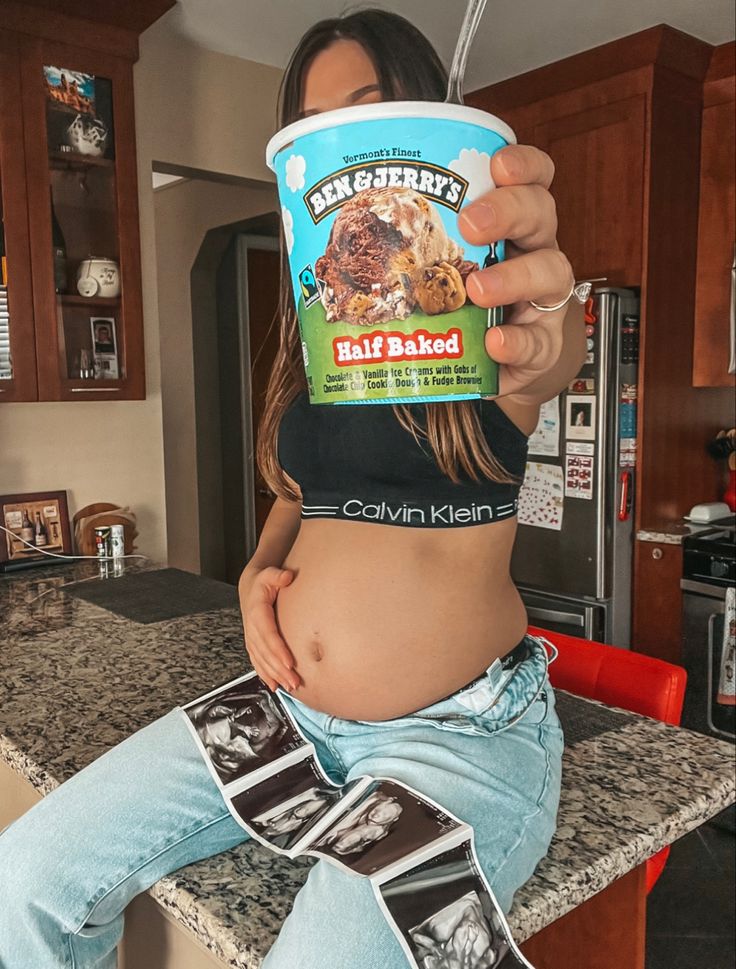 a woman sitting on a kitchen counter holding up a cup of ice cream to her stomach