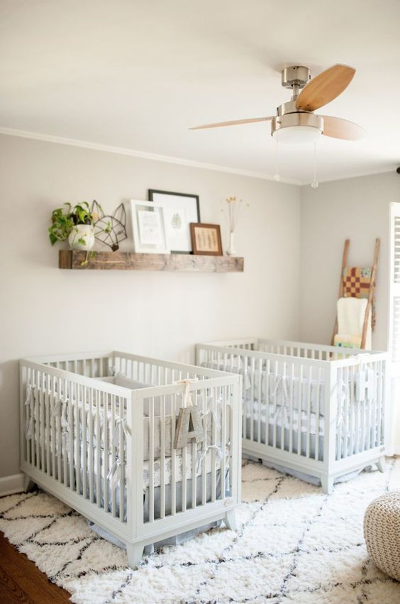 two white cribs sitting in a room with a ceiling fan and pictures on the wall