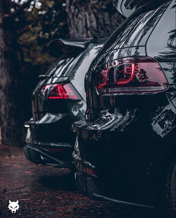 two black cars parked next to each other in the woods with trees behind them and one has its tail lights on