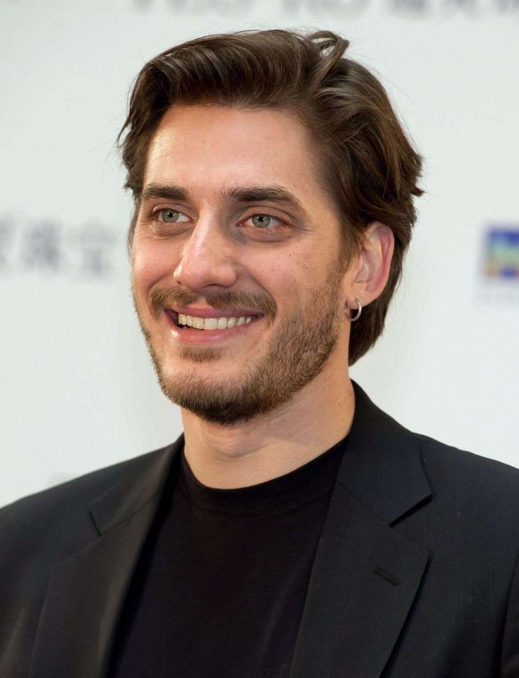 a man in a black suit smiling at the camera with his eyes wide open and hair down