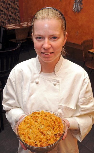 a woman in a chef's uniform holding a dish with cheese on it and looking at the camera