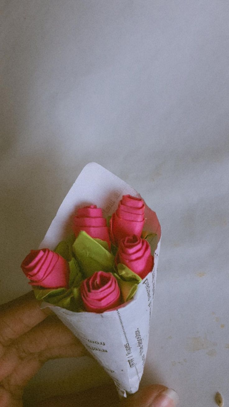 a hand holding a paper cone with pink roses in it and green leaves on top