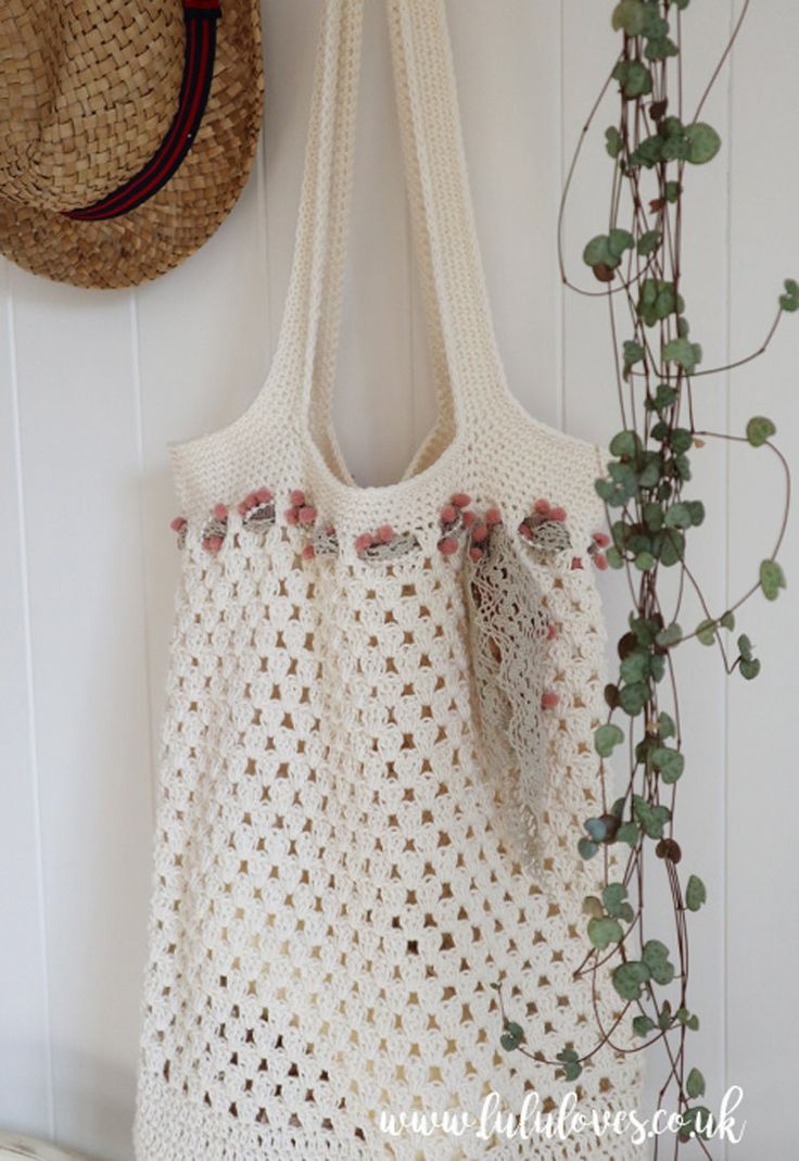 a white crocheted bag hanging on a wall next to a hat and plant